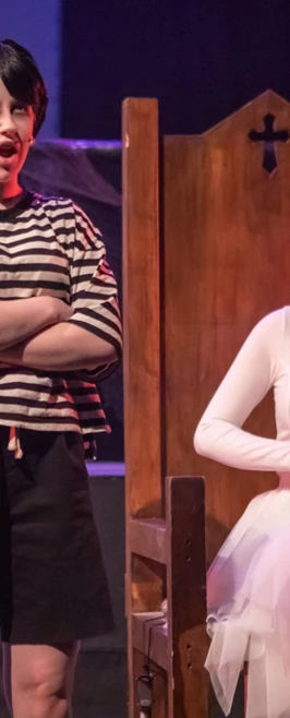 Performer delivering a monologue with a theatrical expression, while another character dressed as a ghost bride sits solemnly nearby Performer delivering a monologue with a theatrical expression, while another character dressed as a ghost bride sits solemnly nearby